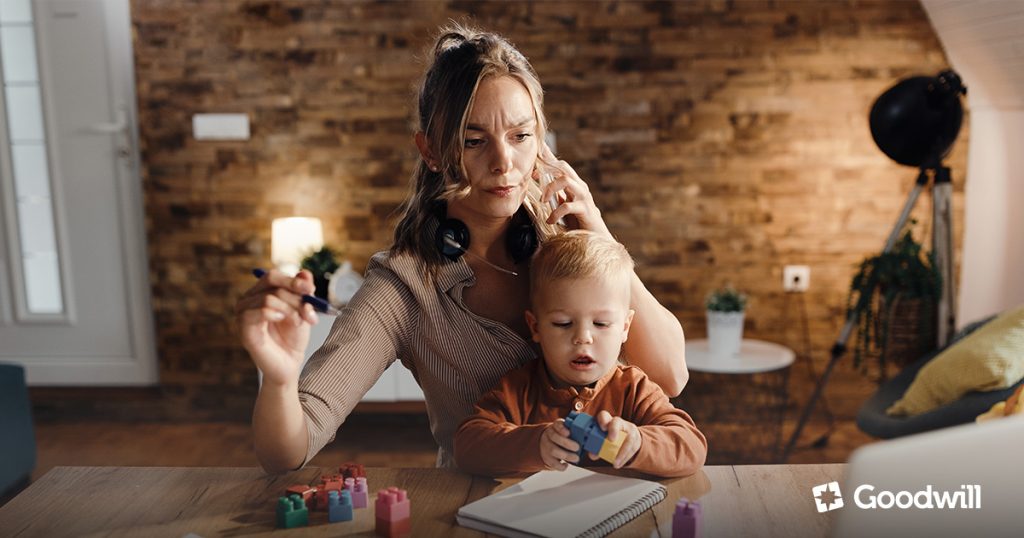 Multitasking anyuka dolgozik otthon laptopon a gyermekével, amely a tartós fizikai és szellemi kimerültség állapotát szimbolizálja.